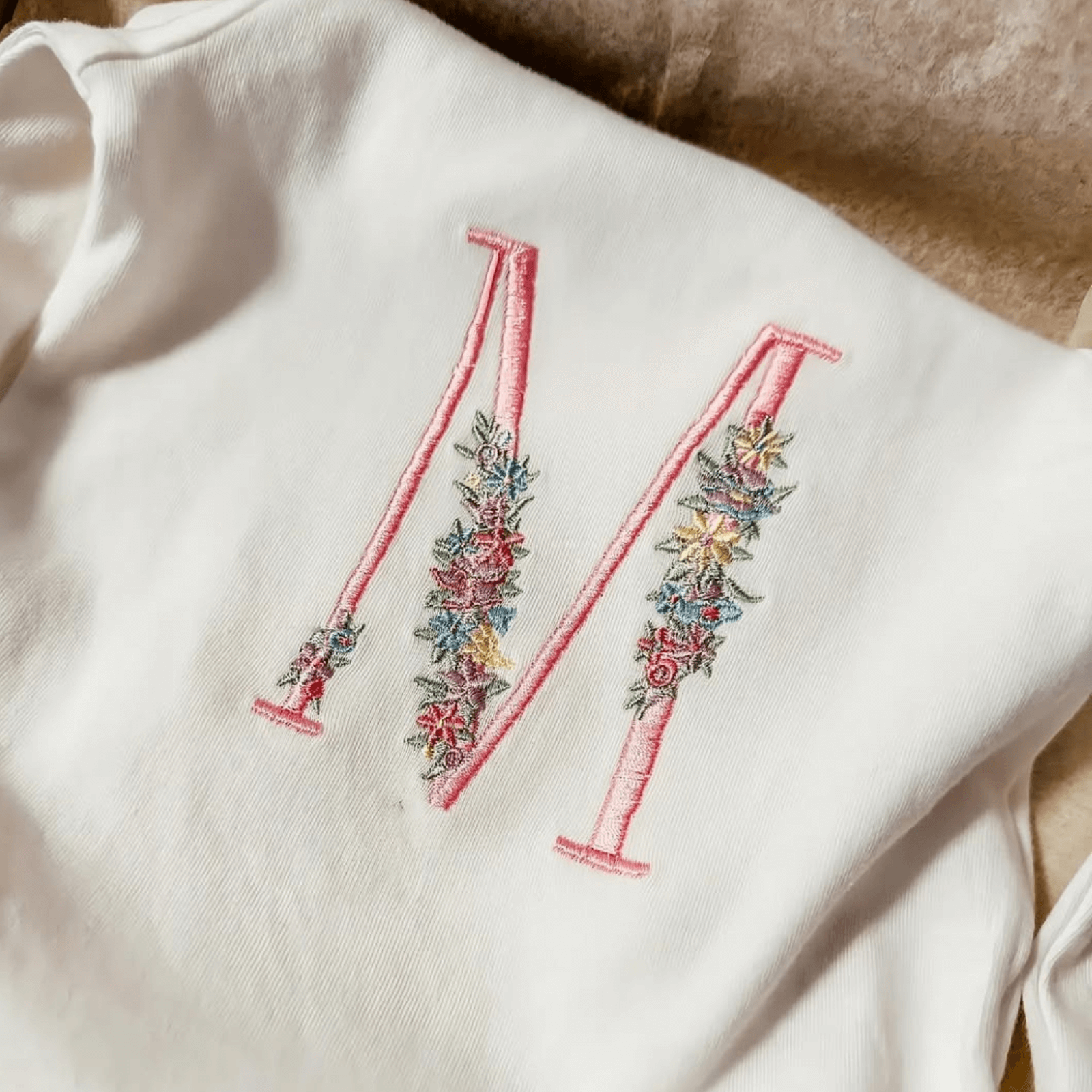 Bílé kojenecké tričko s výraznou a barevnou výšivkou ozdobného písmene M s květinovým designem.