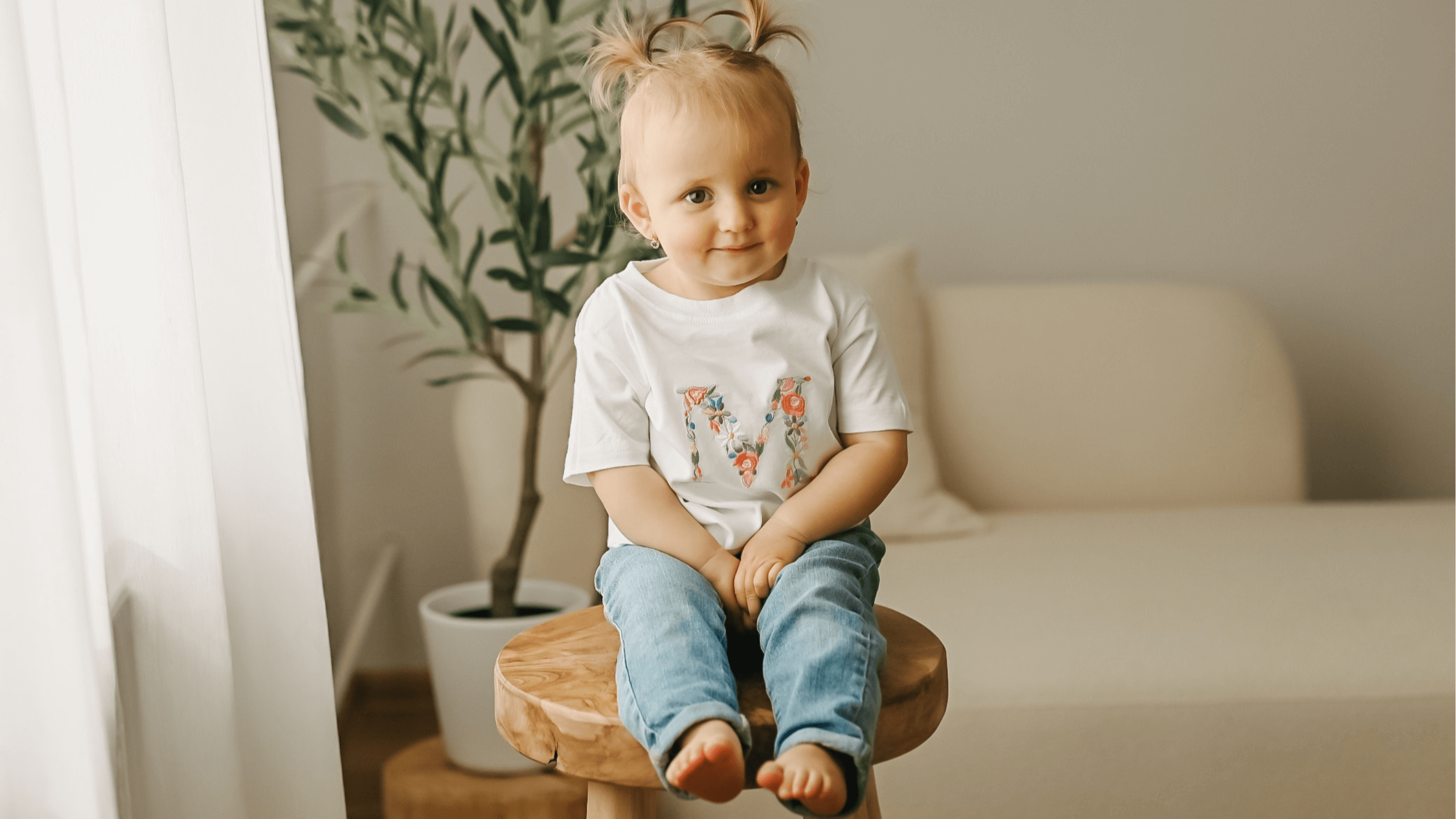 Detailní záběr na roztomilé miminko v bílém tričku s personalizovanou výšivkou velkého písmene M zdobeného růžovými, modrými a žlutými květy.