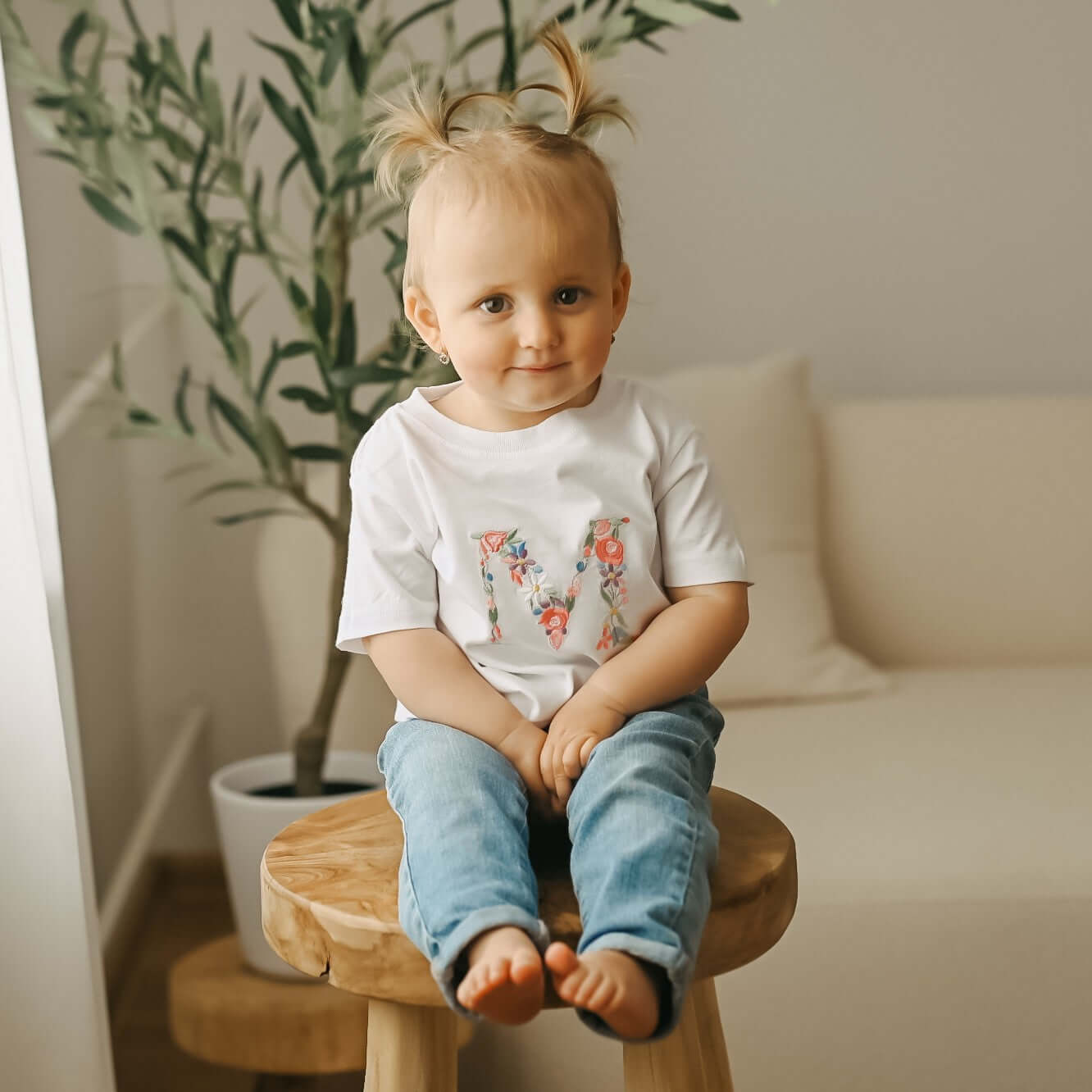 Detailní záběr na roztomilé miminko v bílém tričku s personalizovanou výšivkou velkého písmene M zdobeného růžovými, modrými a žlutými květy.