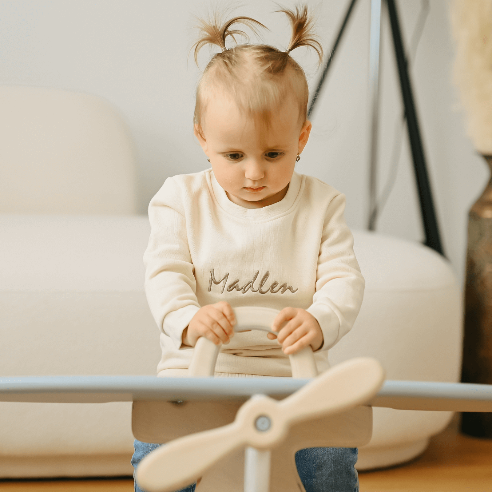 Detailní záběr na hrající si miminko s dřevěným letadlem. Dítě má na sobě personalizovanou mikinou s vyšitým jménem Madlen na hrudi.
