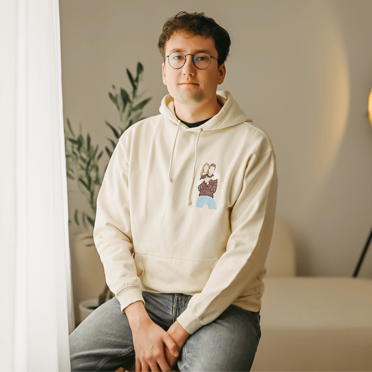 Mladý muž s brýlemi sedí a má na sobě krémovou mikinu s kapucí a barevnou výšivkou siluet dvou lidí na hrudi.