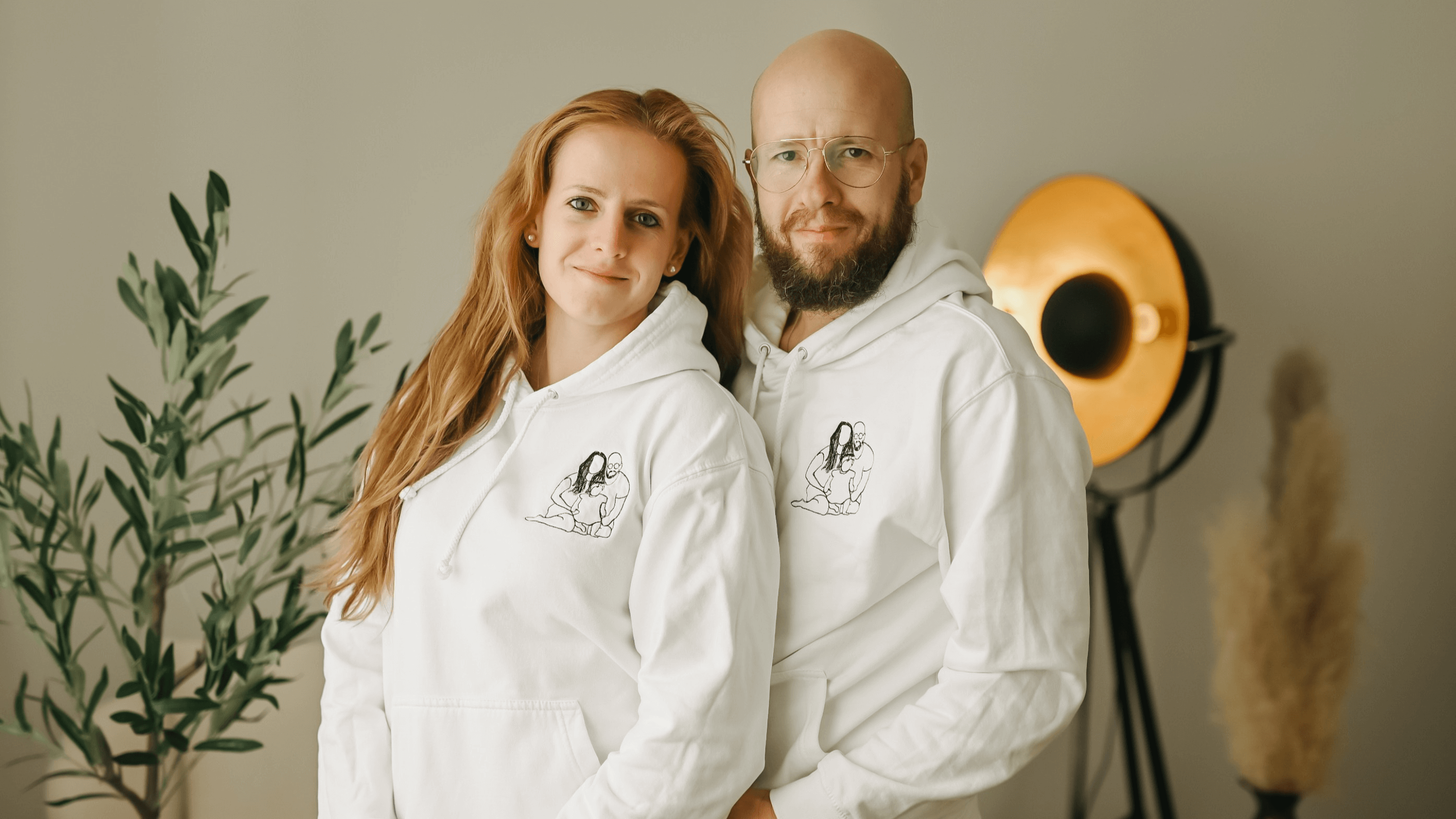 Usmívající se muž a žena v bílých mikinách s kapucí a s černou výšivkou obrysu rodiny s dítětem.