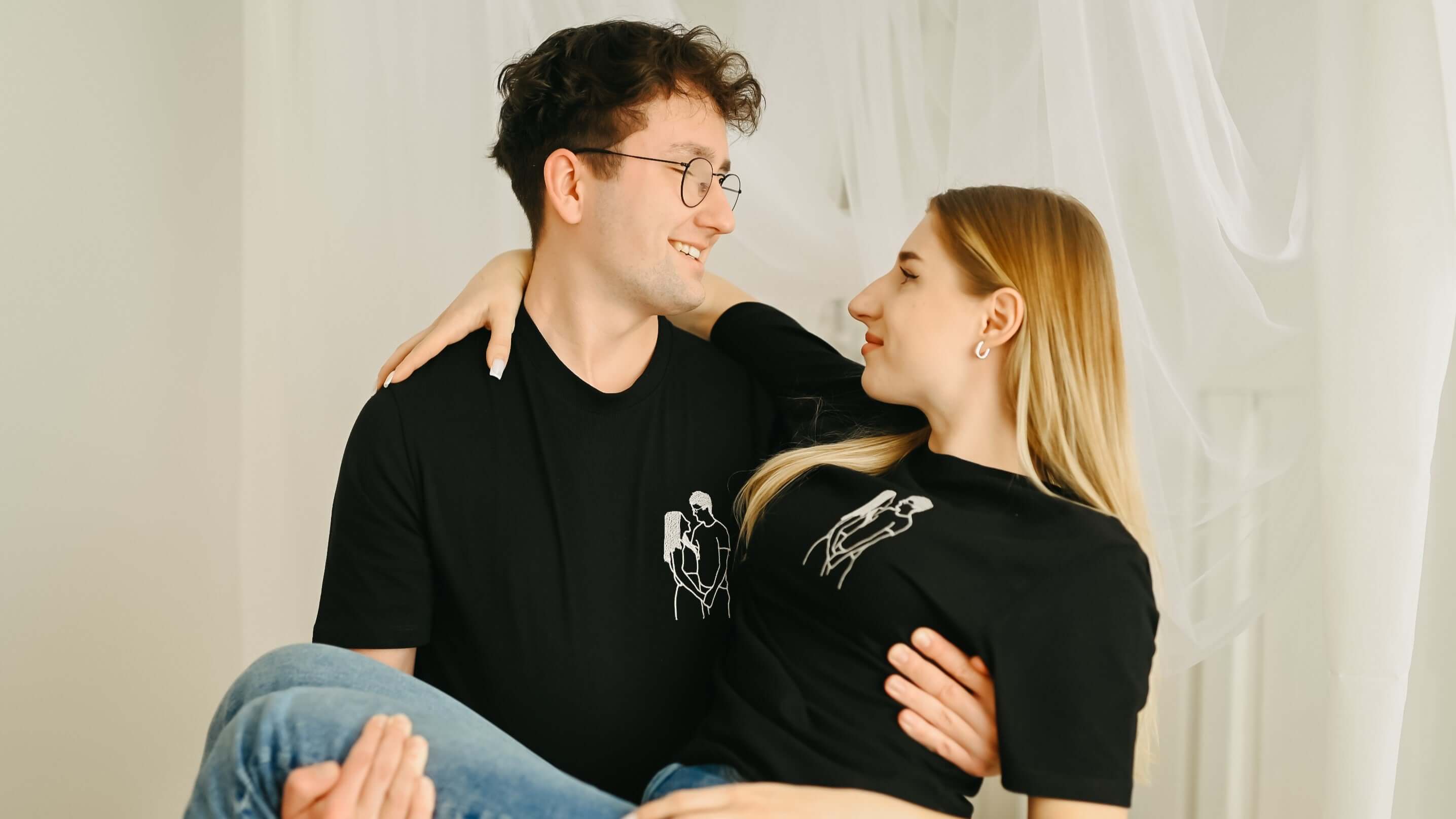 Muž drží ženu a oba mají na sobě černá trička s bílou výšivkou dvou objímajících se lidí.