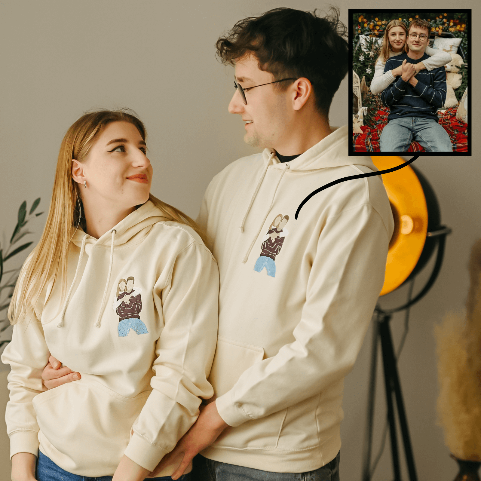 Usmívající se mladý muž s brýlemi a žena v krémových mikinách s kapucí a s vyšitými barevnými siluetami dvou lidí v objetí.