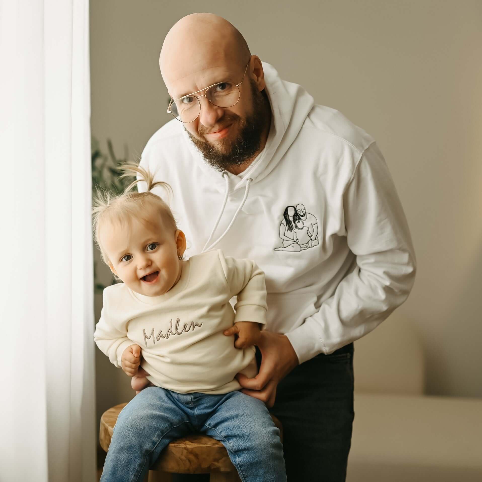 Portrét usměvavého otce držícího svou dceru v náručí. Holčička má na sobě personalizovanou mikinu bez kapuce s vyšitým jménem Madlen.