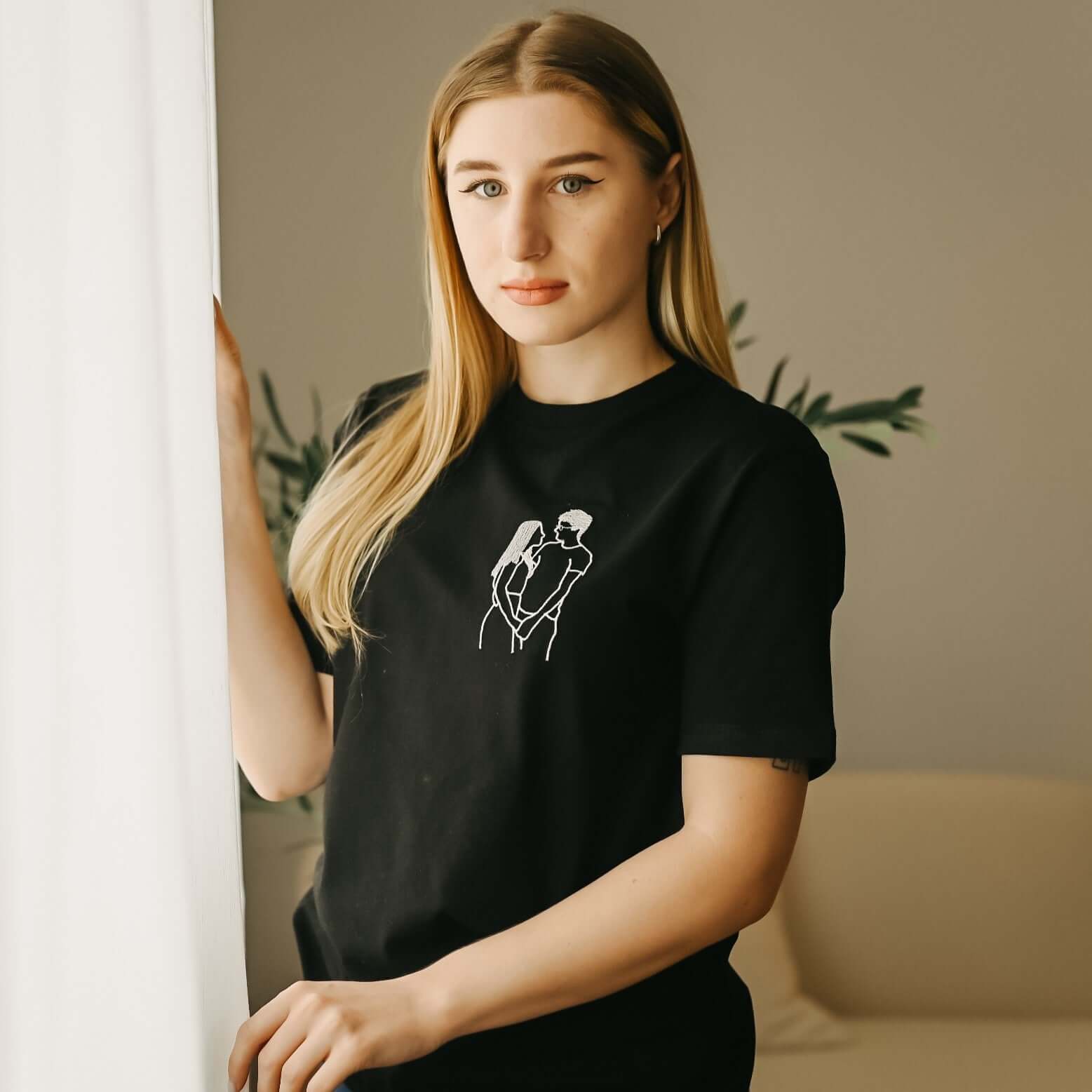 Mladá žena s dlouhými světlými vlasy má na sobě černé tričko s krátkým rukávem a bílou výšivkou siluety objímajícího se páru.